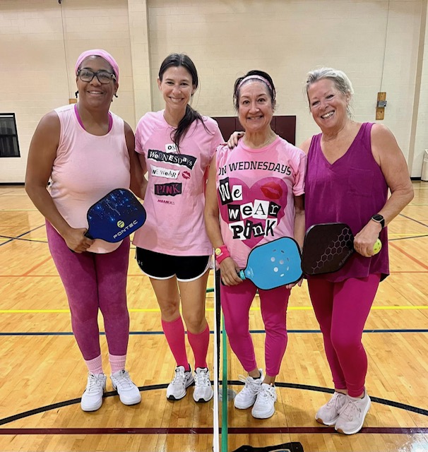 Four women standing on an indoor pickleball court, all dressed in pink and holding their paddles while smiling at the camera.